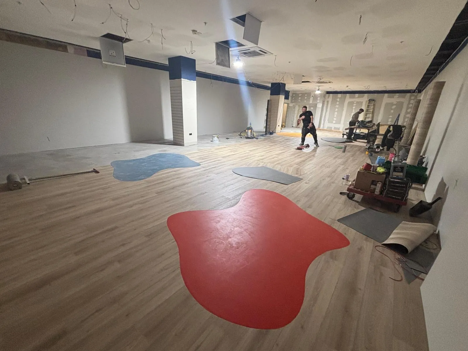 Two fitters at work at the far end of a commercial unit in Uxbridge, with red, blue and grey inlay shapes laid out on a wood-effect floor
