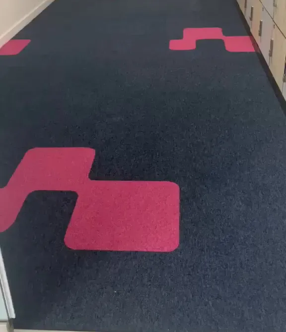 A large pink inlay in the navy carpet tiles of a corridor lined with lockers, with more inlays beyond