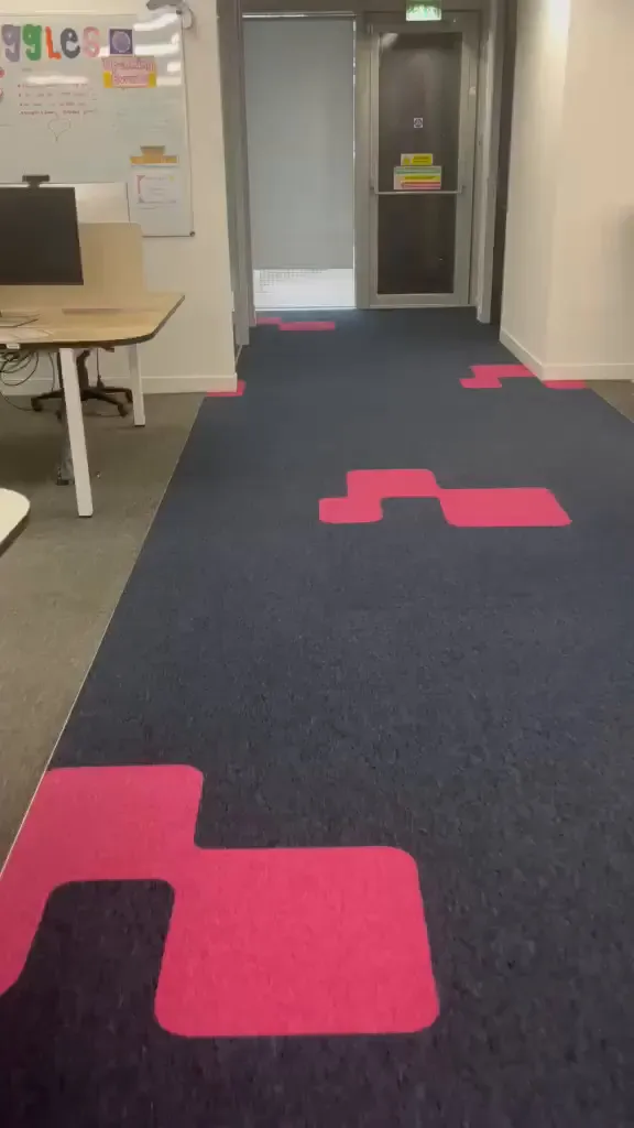 Navy carpet tile walkway with pink inlays leading between desks to a doorway, edged by grey carpet