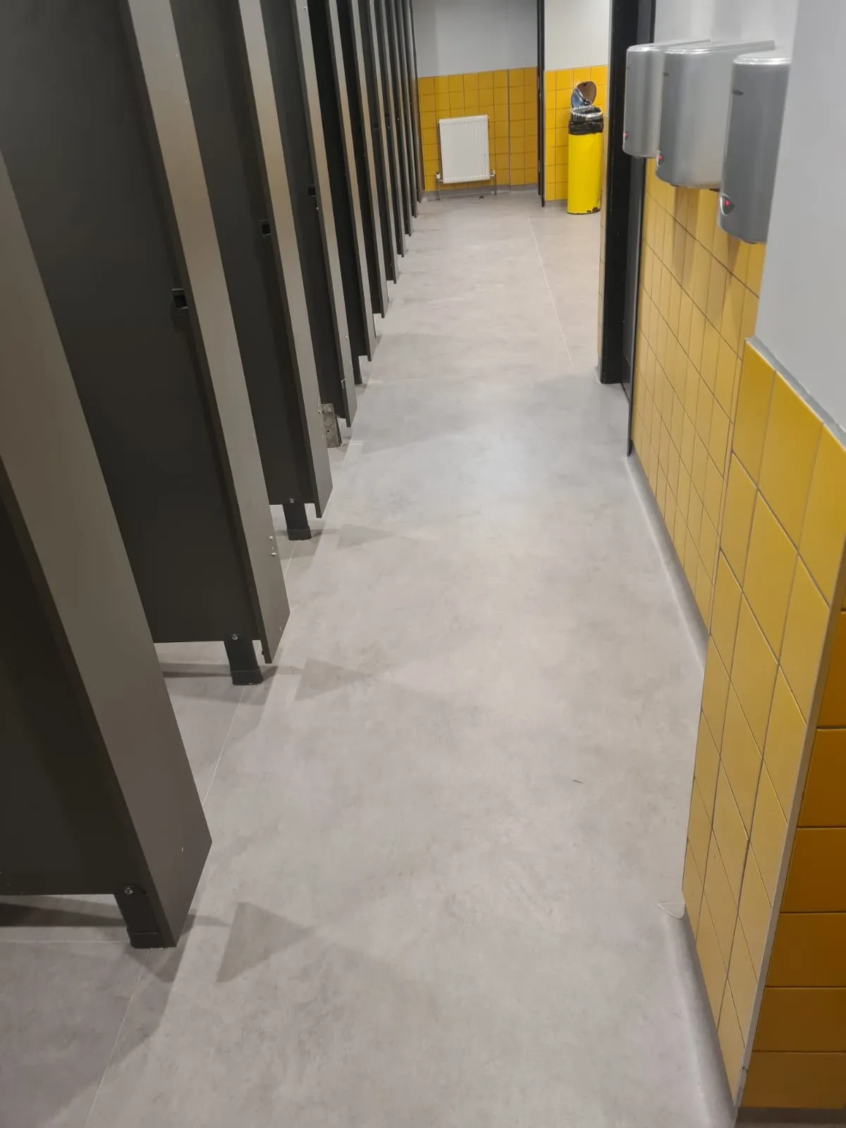 Light grey sheet vinyl running the length of a washroom, past a row of full-height cubicles and a tiled wall